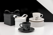 Zwei Espresso-Tassen mit Untertassen in Schwarz und Weiß mit Disney-Motiv, arrangiert neben Geschenkboxen; Perspektive: leicht seitliche Frontansicht auf Tischhöhe.