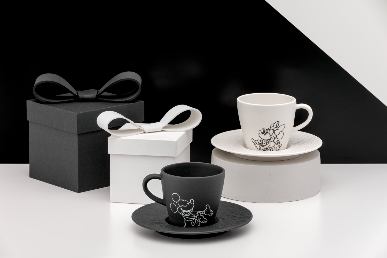 Zwei Espresso-Tassen mit Untertassen in Schwarz und Weiß mit Disney-Motiv, arrangiert neben Geschenkboxen Perspektive: leicht seitliche Frontansicht auf Tischhöhe.