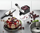 Kirschentkerner CORER in der Küche, gefüllt mit Kirschen, neben einem Teller mit einem Kirschdessert und einer Schüssel mit Sahne, aus der Vogelperspektive.