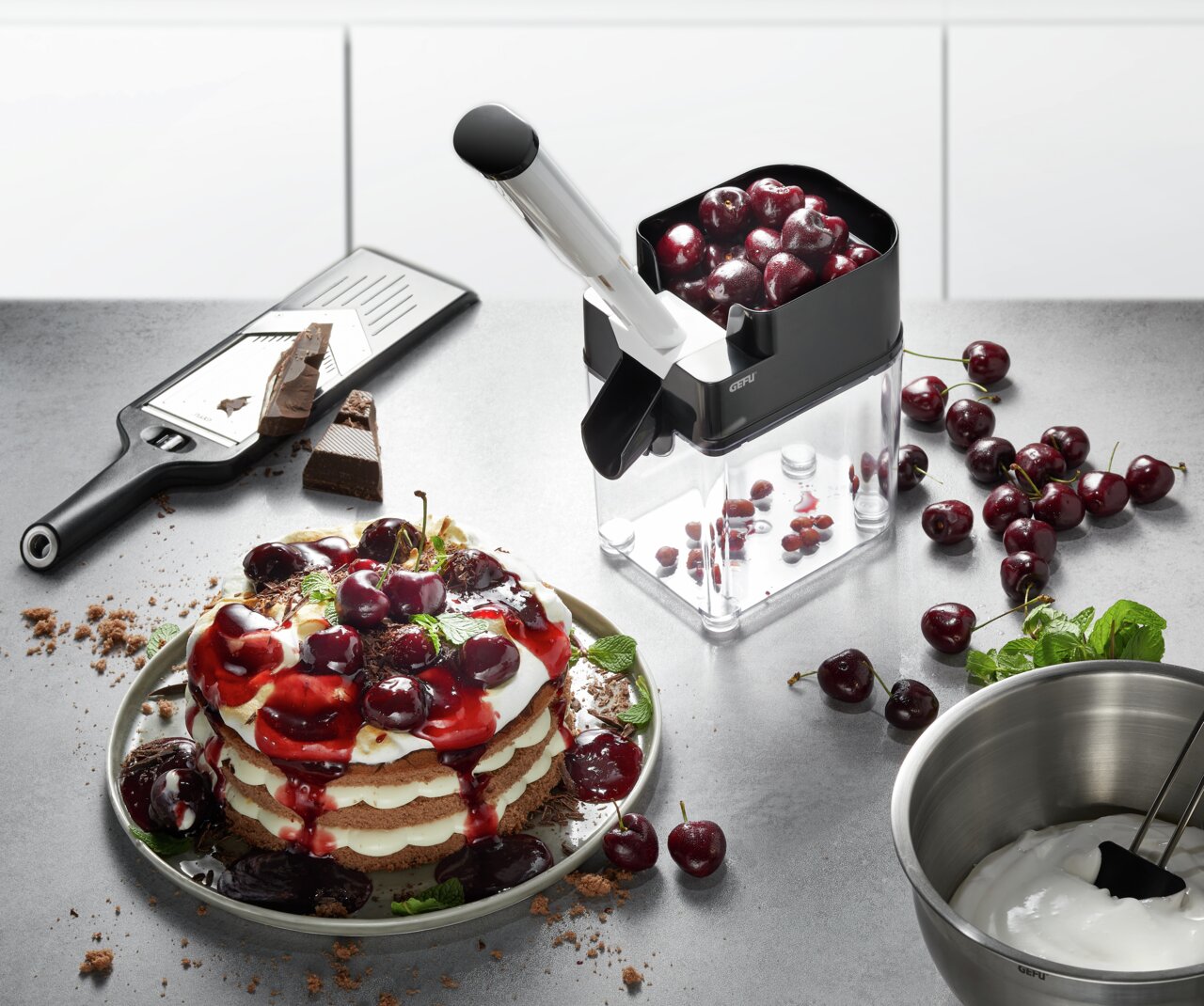 GEFU Kersenontpitter CORER Een CORER-kersenontpitter in de keuken, gevuld met kersen, naast een bord met een kersendessert en een schaal met slagroom, gezien vanuit vogelperspectief.