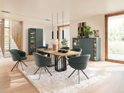 Modern ingerichte eetkamer met een houten eettafel, zes groen gestoffeerde kuipstoelen en bijpassende donkergroene kasten, gezien vanuit schuin vooraanzicht.