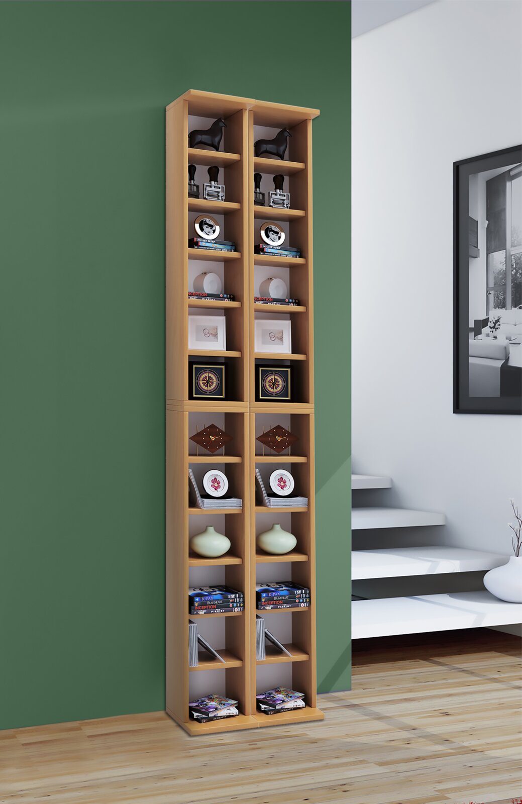 Hoher, schmaler Bücher- und CD-Regal-Turm aus hellem Holz mit vielen offenen Fächern, dekoriert mit Büchern, DVDs und Dekoobjekten, vor grüner Wand im Wohnraum, Frontalansicht in leichter Perspektive von rechts.