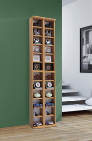 Hoher, schmaler Bücher- und CD-Regal-Turm aus hellem Holz mit vielen offenen Fächern, dekoriert mit Büchern, DVDs und Dekoobjekten, vor grüner Wand im Wohnraum, Frontalansicht in leichter Perspektive von rechts.