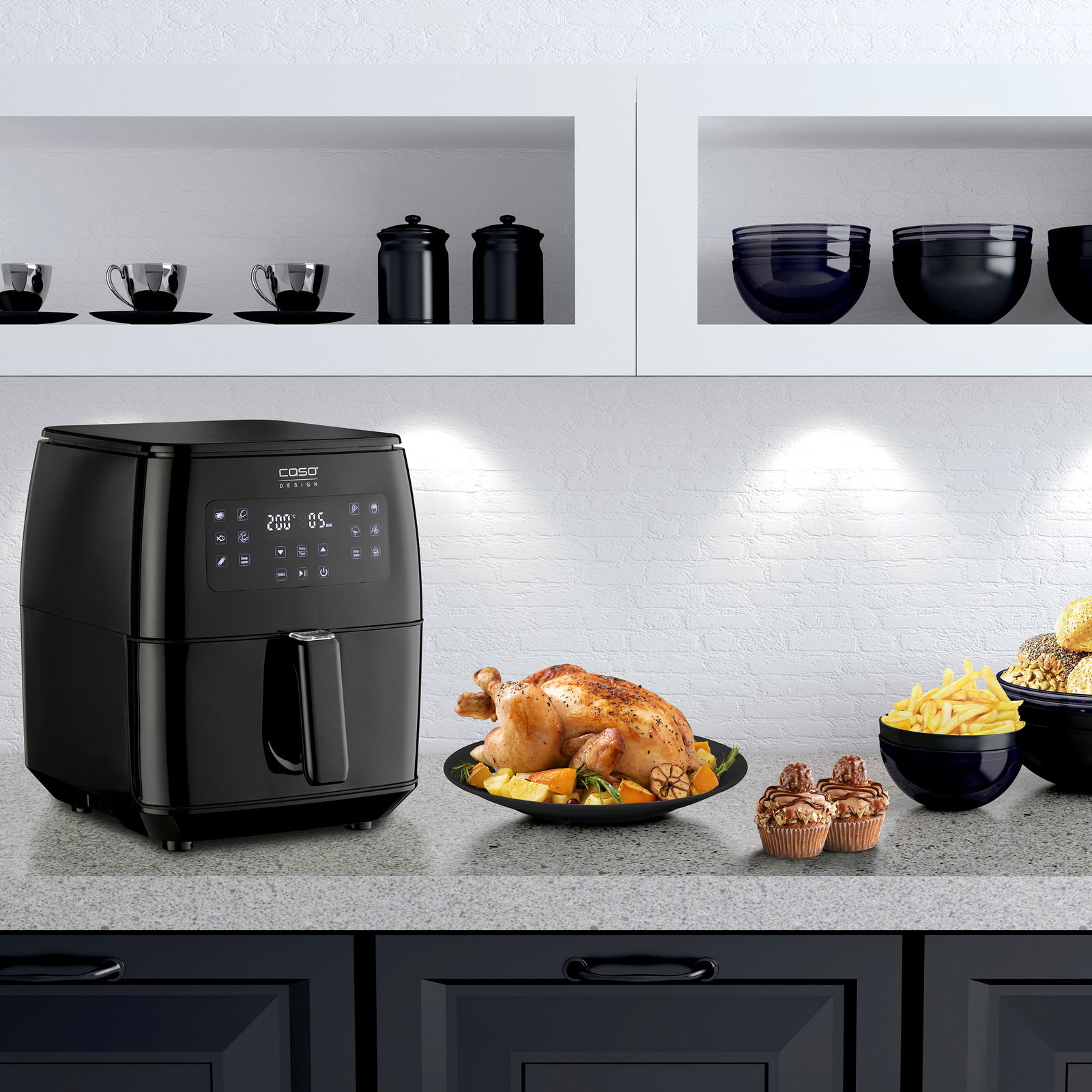 Schwarze Heißluftfritteuse auf einer Küchenarbeitsplatte, umgeben von einem gebratenen Hähnchen, Pommes frites und Muffins, aus einer frontalen Perspektive.