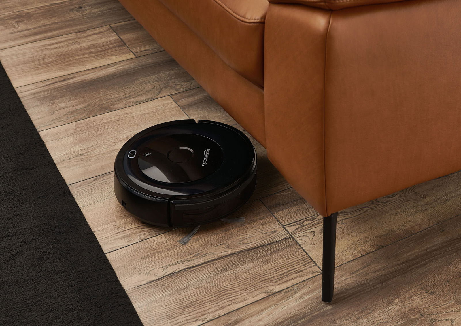 Ein schwarzer Saugroboter reinigt den Holzboden unter einem braunen Sofa aus der Seitenperspektive.