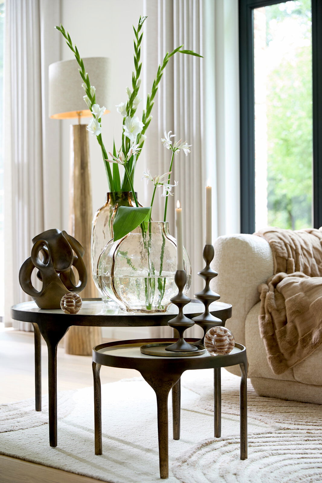 Zwei runde Beistelltische mit dekorativen Objekten, darunter eine abstrakte Skulptur, Glasvasen mit Blumen und Kerzenhalter, in einem modernen Wohnzimmer mit natürlichem Licht von der Seite.