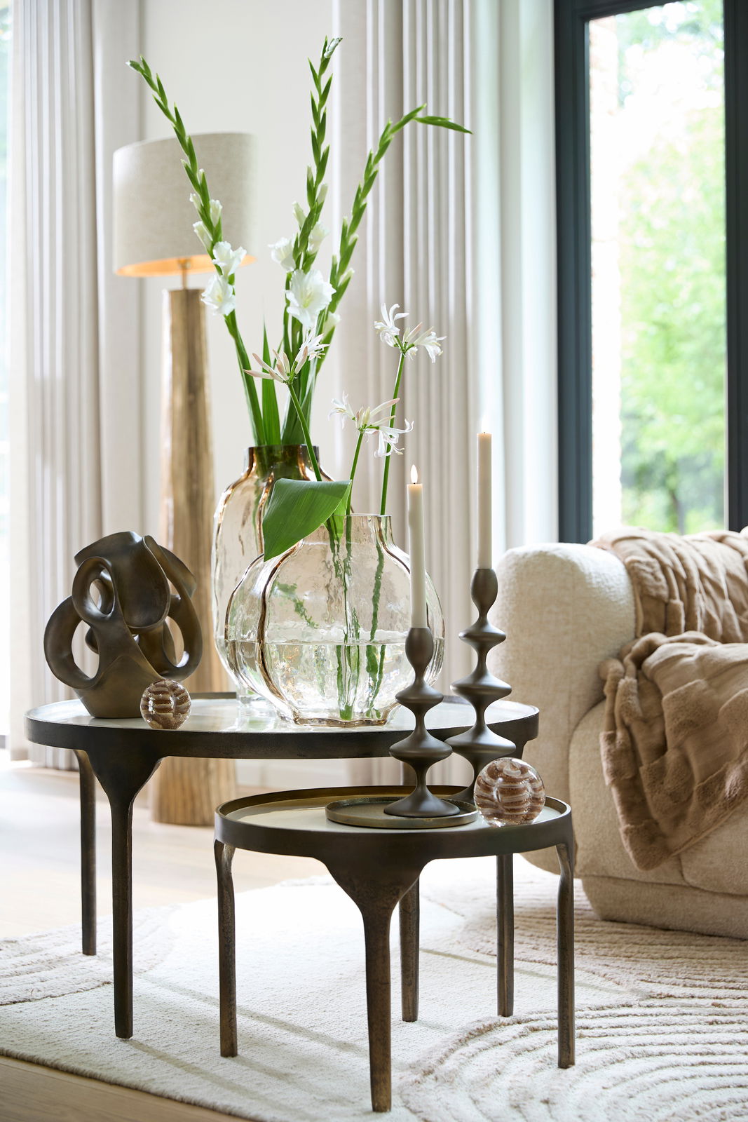 Light&Living Ornament THALINA Zwei runde Beistelltische mit dekorativen Objekten, darunter eine abstrakte Skulptur, Glasvasen mit Blumen und Kerzenhalter, in einem modernen Wohnzimmer mit natürlichem Licht von der Seite.