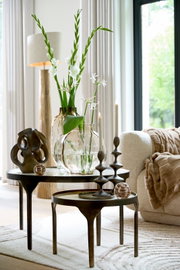 Twee ronde bijzettafeltjes met decoratieve voorwerpen, waaronder een abstract beeld, glazen vazen met bloemen en kandelaars, in een moderne woonkamer met natuurlijk licht dat van opzij binnenvalt.