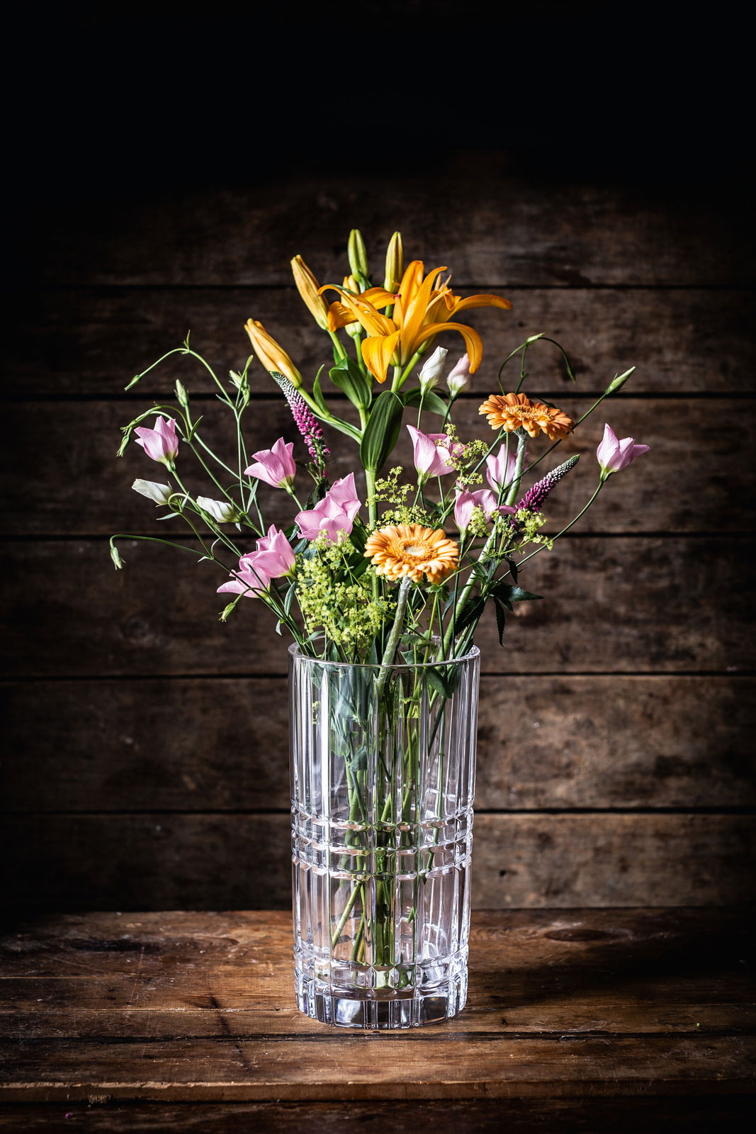 Klare, quadratische Vase mit Blumenstrauß aus orangefarbenen Lilien und rosa Blüten, frontal auf Holztisch vor Holzwand.