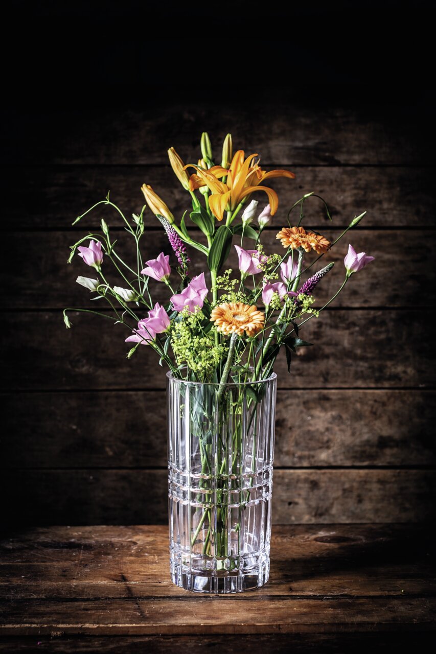 Klare, quadratische Vase mit Blumenstrauß aus orangefarbenen Lilien und rosa Blüten, frontal auf Holztisch vor Holzwand.