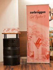 Roze ‘Zurbrüggen Get Together’-eventbanner naast een zwarte statafel met feestelijk gedecoreerde glazen.
