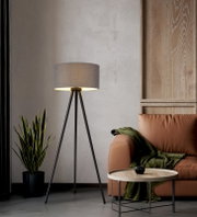 Moderne Dreibein-Stehlampe mit schwarzem Metallgestell und grauem Stoffschirm, eingeschaltet im Wohnzimmer neben Ledersofa und Beistelltisch, frontale Perspektive