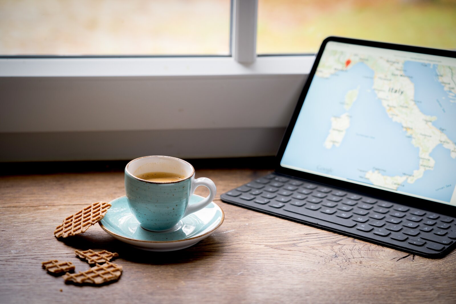 Türkise Kaffeetasse mit Espresso auf Untertasse neben Waffelkeksen auf Holztisch, rechts ein Tablet mit Tastatur und Italien-Karte am Fenster; Perspektive: seitliche Tischansicht in leichtem Schrägwinkel von vorn.