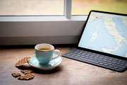 Türkise Kaffeetasse mit Espresso auf Untertasse neben Waffelkeksen auf Holztisch, rechts ein Tablet mit Tastatur und Italien-Karte am Fenster; Perspektive: seitliche Tischansicht in leichtem Schrägwinkel von vorn.