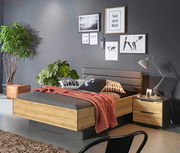 Modernes Schlafzimmer mit einem Bett mit Sockelschubkasten in Holzoptik, seitliche Perspektive. Das Bett ist mit grauer Bettwäsche und einem roten Überwurf dekoriert, umgeben von Pflanzen und stilvollen Wandbildern.