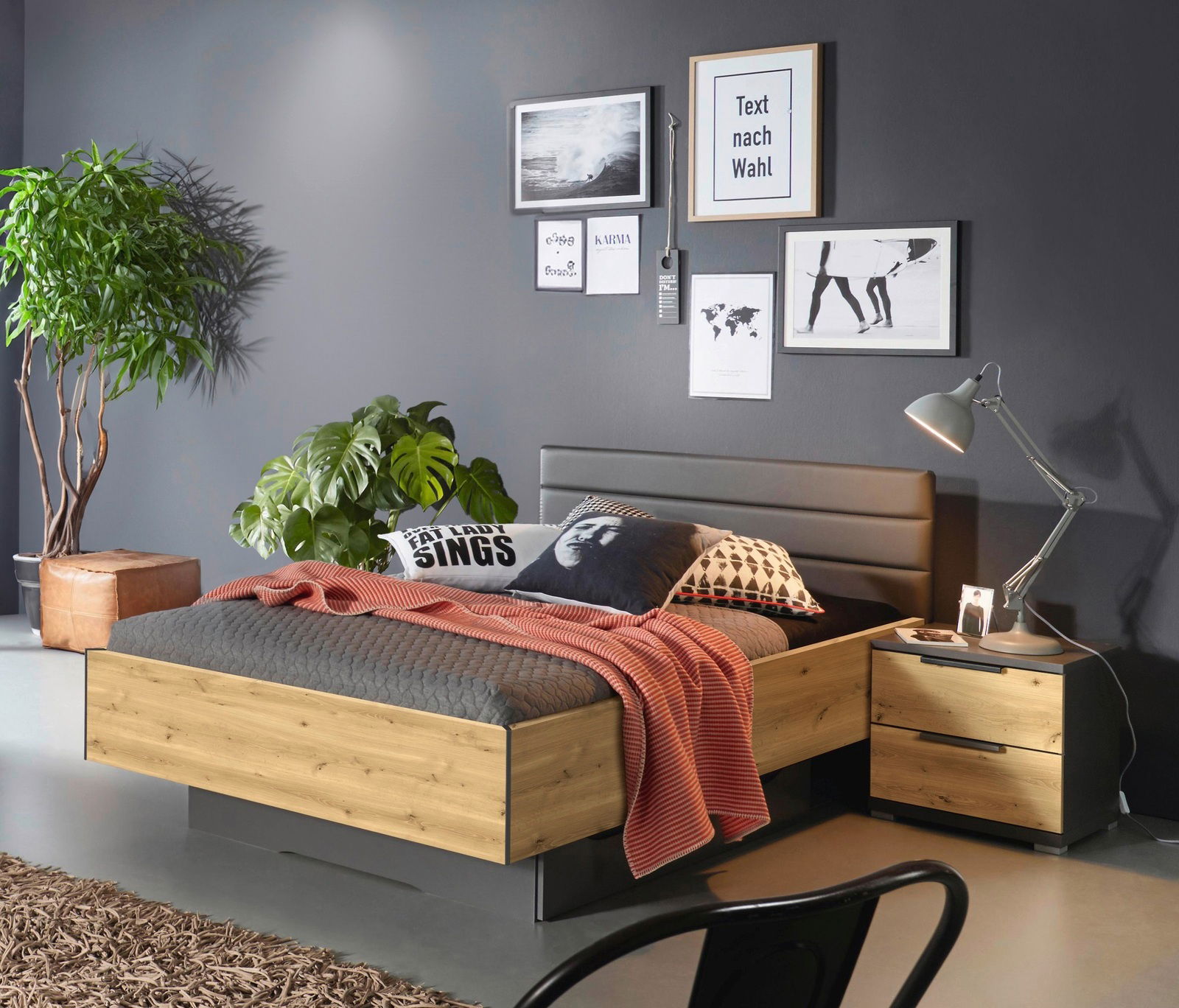 Modernes Schlafzimmer mit einem Bett mit Sockelschubkasten in Holzoptik, seitliche Perspektive. Das Bett ist mit grauer Bettwäsche und einem roten Überwurf dekoriert, umgeben von Pflanzen und stilvollen Wandbildern.