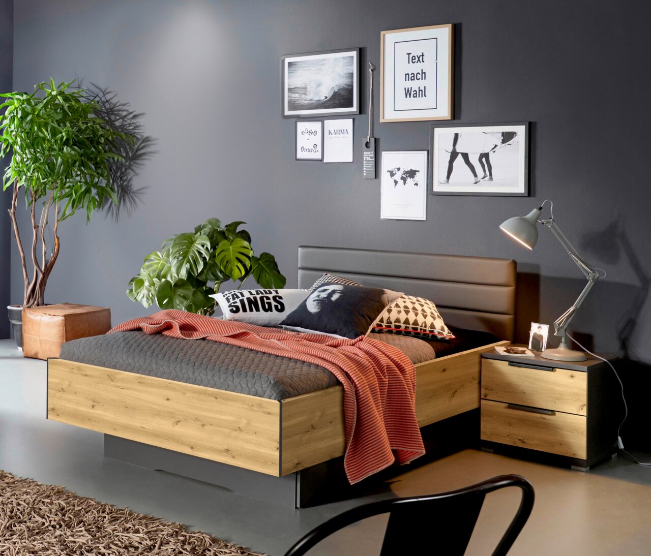 Modernes Schlafzimmer mit einem Bett mit Sockelschubkasten in Holzoptik, seitliche Perspektive. Das Bett ist mit grauer Bettwäsche und einem roten Überwurf dekoriert, umgeben von Pflanzen und stilvollen Wandbildern.