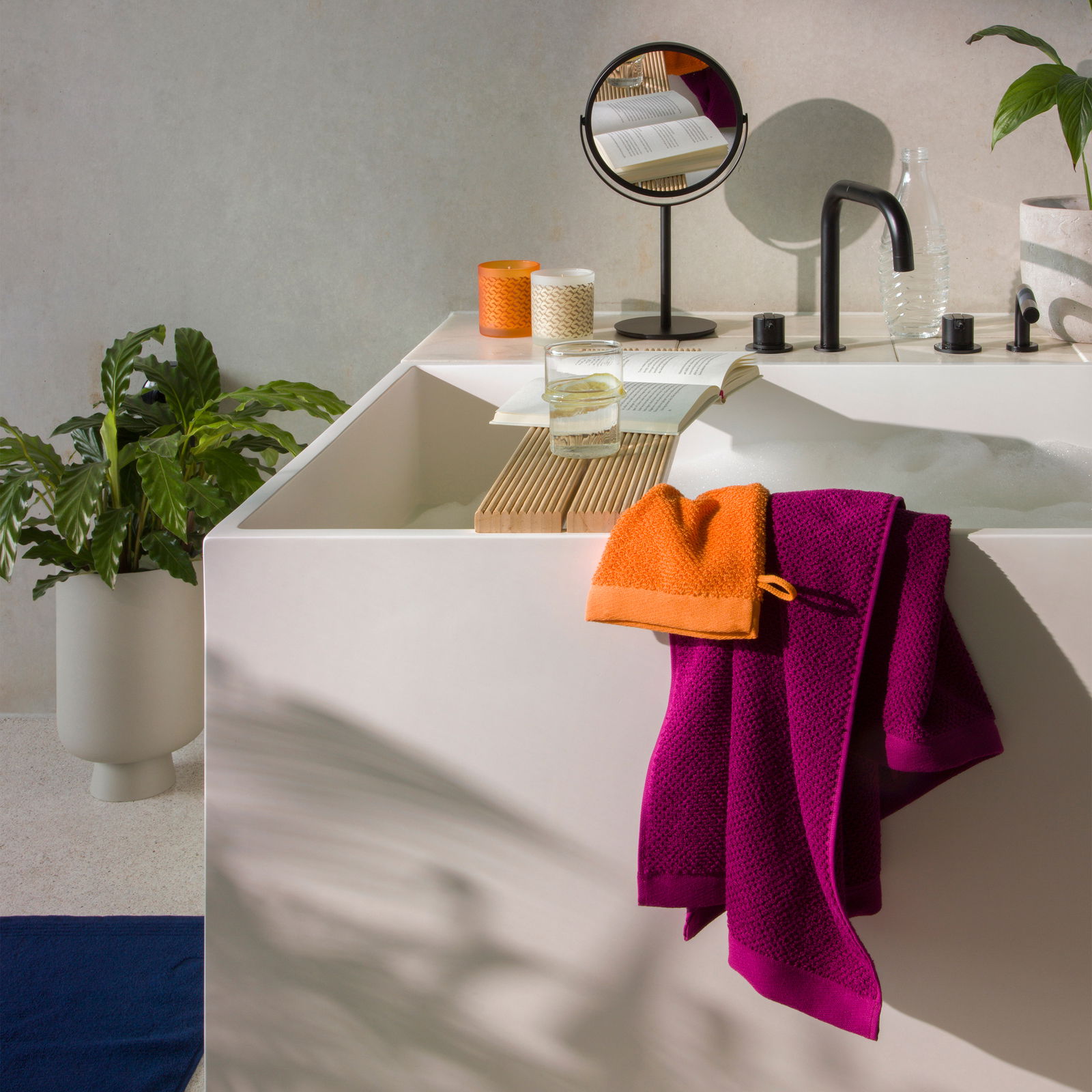 Blick auf ein modernes Badezimmer mit einem weißen Waschbecken, auf dem ein lila und ein orangefarbenes Handtuch liegen. Im Hintergrund sind ein Spiegel, ein Buch, ein Glas Wasser und Pflanzen zu sehen.