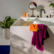 Blick auf ein modernes Badezimmer mit einem weißen Waschbecken, auf dem ein lila und ein orangefarbenes Handtuch liegen. Im Hintergrund sind ein Spiegel, ein Buch, ein Glas Wasser und Pflanzen zu sehen.