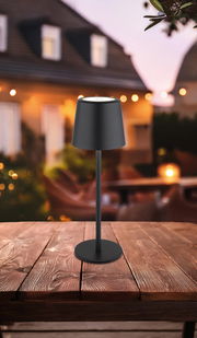 Moderne schwarze Tischleuchte auf einem Holztisch, im Freien bei Dämmerung fotografiert.