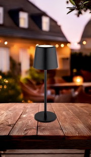 Moderne schwarze Tischleuchte auf einem Holztisch, im Freien bei Dämmerung fotografiert.