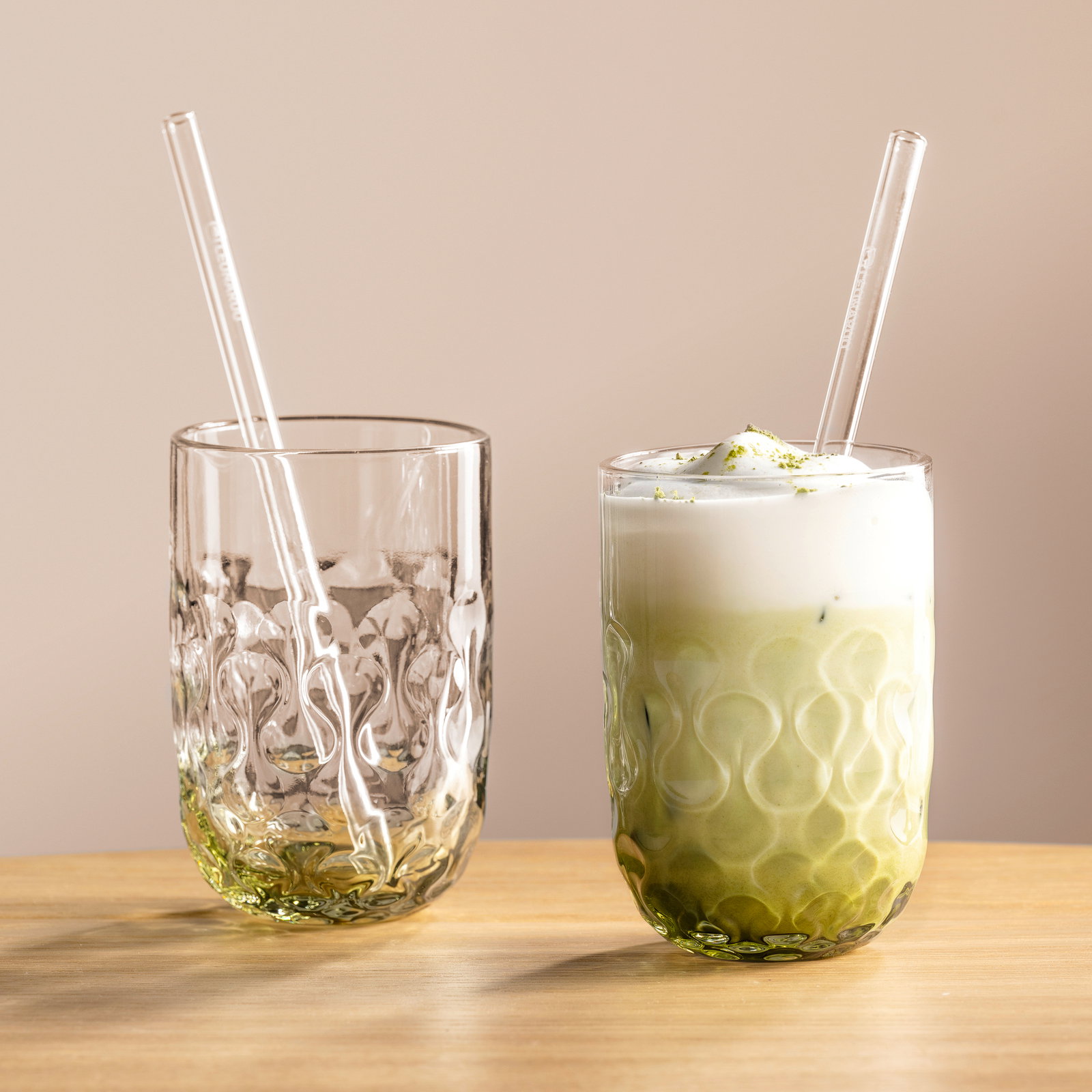 Zwei elegante Gläser mit strukturiertem Design, eines gefüllt mit Matcha Latte und das andere leer, beide mit Glasstrohhalmen, auf einem Holztisch vor neutralem Hintergrund, seitliche Perspektive.