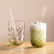 Zwei elegante Gläser mit strukturiertem Design, eines gefüllt mit Matcha Latte und das andere leer, beide mit Glasstrohhalmen, auf einem Holztisch vor neutralem Hintergrund, seitliche Perspektive.
