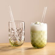 Zwei elegante Gläser mit strukturiertem Design, eines gefüllt mit Matcha Latte und das andere leer, beide mit Glasstrohhalmen, auf einem Holztisch vor neutralem Hintergrund, seitliche Perspektive.