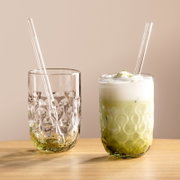 Twee elegante glazen met een gestructureerd ontwerp, het ene gevuld met matcha latte en het andere leeg, beide met glazen rietjes, op een houten tafel tegen een neutrale achtergrond, gezien vanuit een zijdelingse hoek.