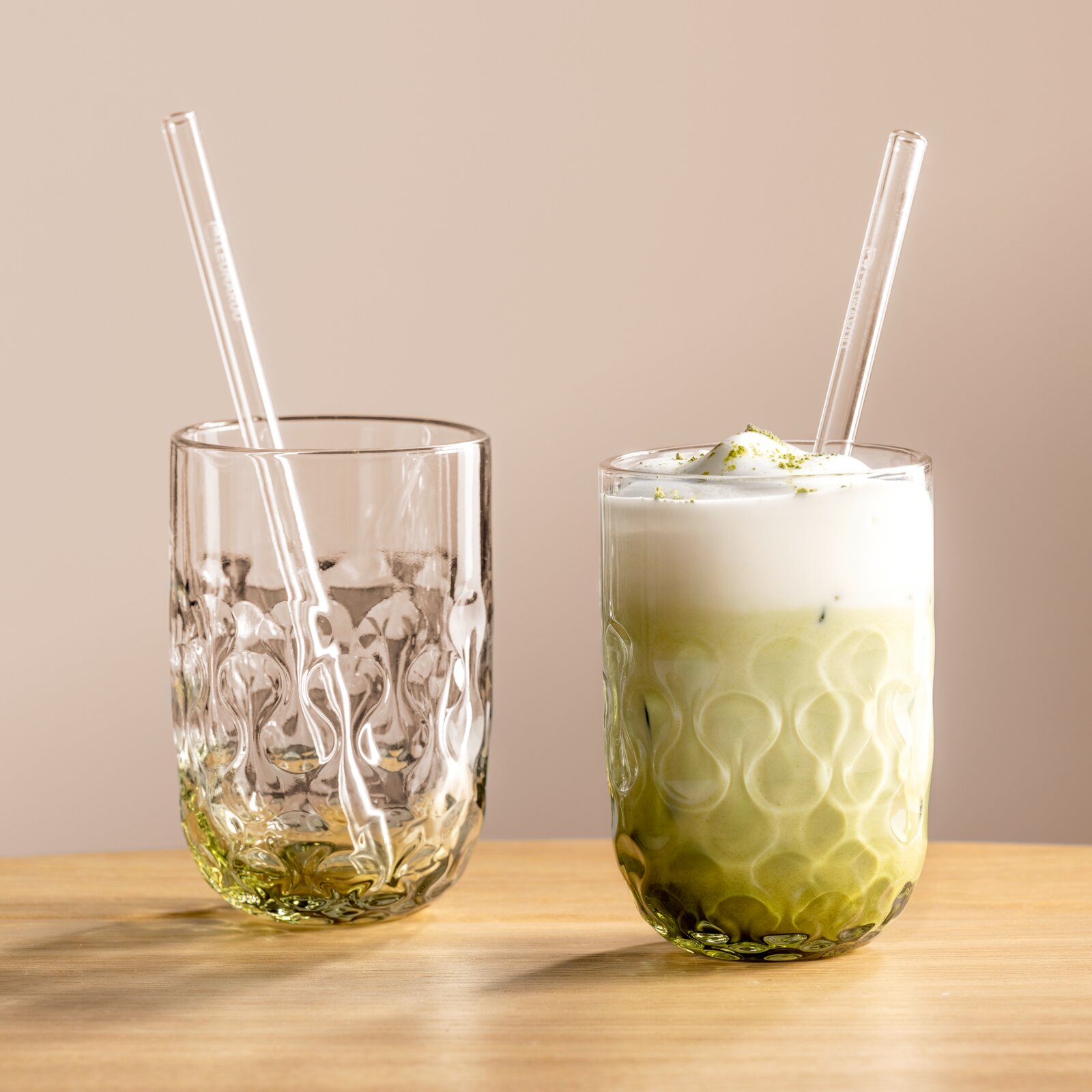 Twee elegante glazen met een gestructureerd ontwerp, het ene gevuld met matcha latte en het andere leeg, beide met glazen rietjes, op een houten tafel tegen een neutrale achtergrond, gezien vanuit een zijdelingse hoek.