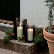 Zwei schwarze Metall-Laternen mit Glaszylinder und brennenden weißen Stumpenkerzen auf einer Steinstufe vor einer Hauswand, dekoriert mit Tannenzweigen und roten Beeren, perspektivisch in leichter Seitenansicht auf Augenhöhe