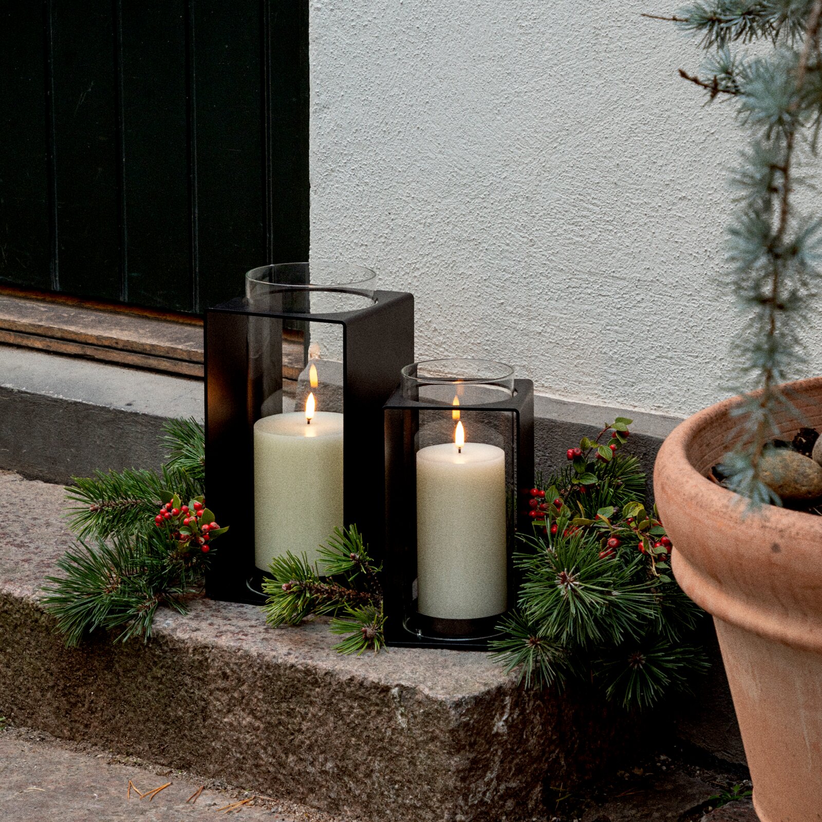 Zwei schwarze Metall-Laternen mit Glaszylinder und brennenden weißen Stumpenkerzen auf einer Steinstufe vor einer Hauswand, dekoriert mit Tannenzweigen und roten Beeren, perspektivisch in leichter Seitenansicht auf Augenhöhe