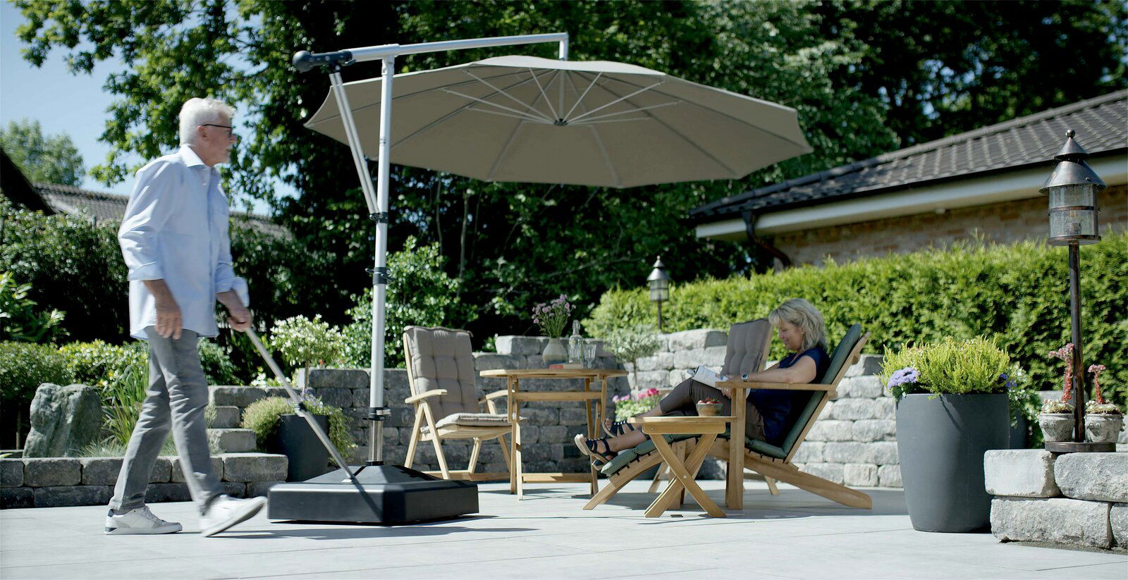 Moderner Schirmständer auf einer Terrasse, mit einem großen Sonnenschirm, der Schatten auf eine Sitzgruppe mit zwei Personen wirft. Perspektive von der Seite.