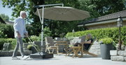 Moderner Schirmständer auf einer Terrasse, mit einem großen Sonnenschirm, der Schatten auf eine Sitzgruppe mit zwei Personen wirft. Perspektive von der Seite.
