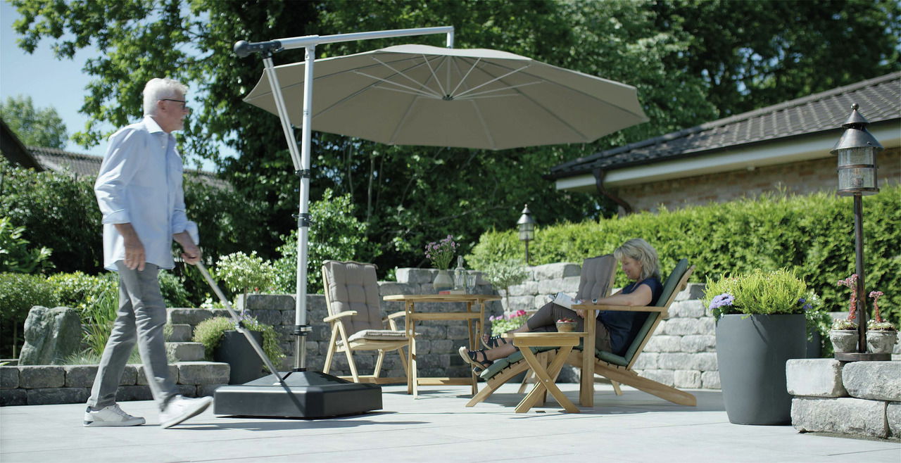 Liro Schirmständer MIDI PLUS Moderner Schirmständer auf einer Terrasse, mit einem großen Sonnenschirm, der Schatten auf eine Sitzgruppe mit zwei Personen wirft. Perspektive von der Seite.