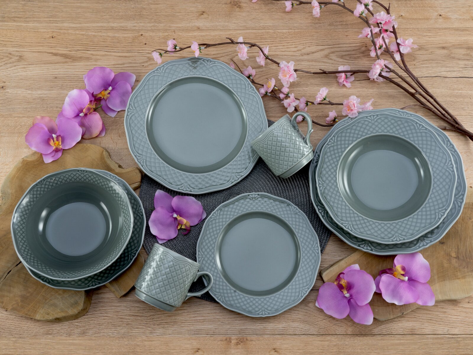 Grau-blaues Keramik-Geschirrset mit Tellern, Schalen und Tassen in Relief-Optik auf Holztisch mit Blumen dekoriert, Draufsicht