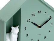 Grüne Wanduhr mit schwarzem Zifferblatt und Zeigern, dekoriert mit einer weißen Katzenfigur in einem seitlichen Fach, seitliche Perspektive.