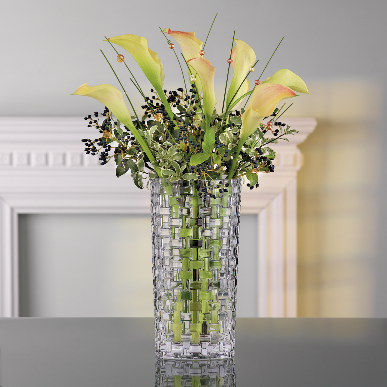 Glasvase mit gewebtem Muster, gefüllt mit gelben Calla-Lilien und grünen Blättern, auf einem Tisch vor einem dekorativen Rahmen, seitliche Perspektive.