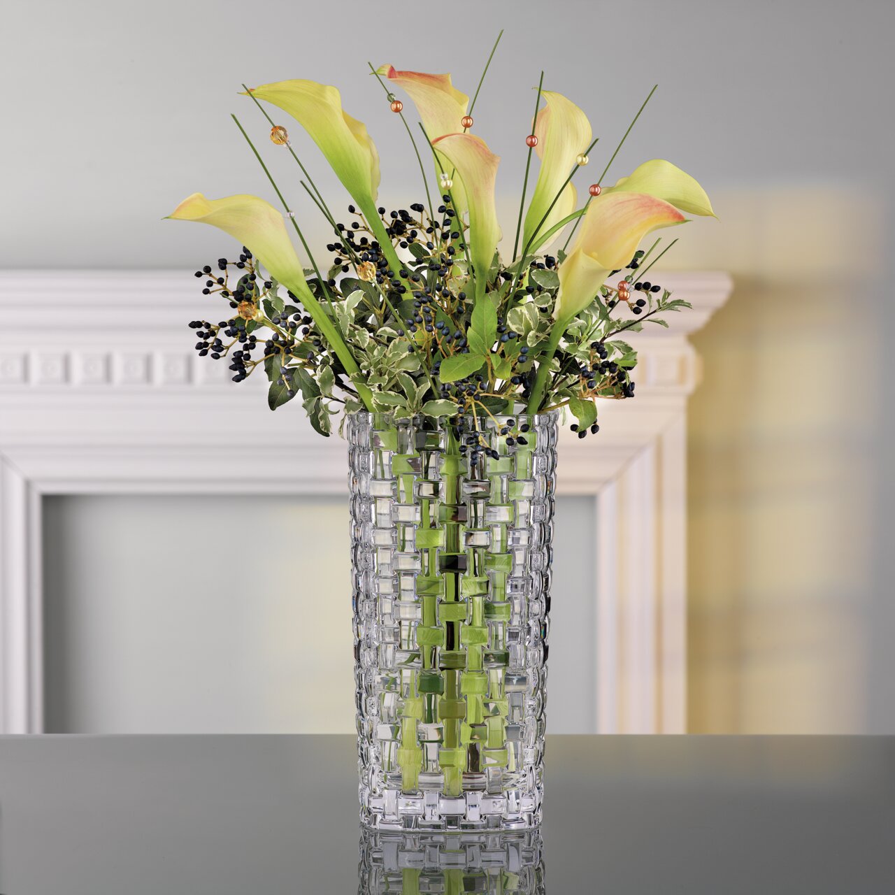 Glasvase mit gewebtem Muster, gefüllt mit gelben Calla-Lilien und grünen Blättern, auf einem Tisch vor einem dekorativen Rahmen, seitliche Perspektive.