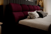 Zijaanzicht van een elegant gestoffeerd bed met een wijnrood hoofdeinde en diverse kussens in een stijlvol ingerichte slaapkamer.