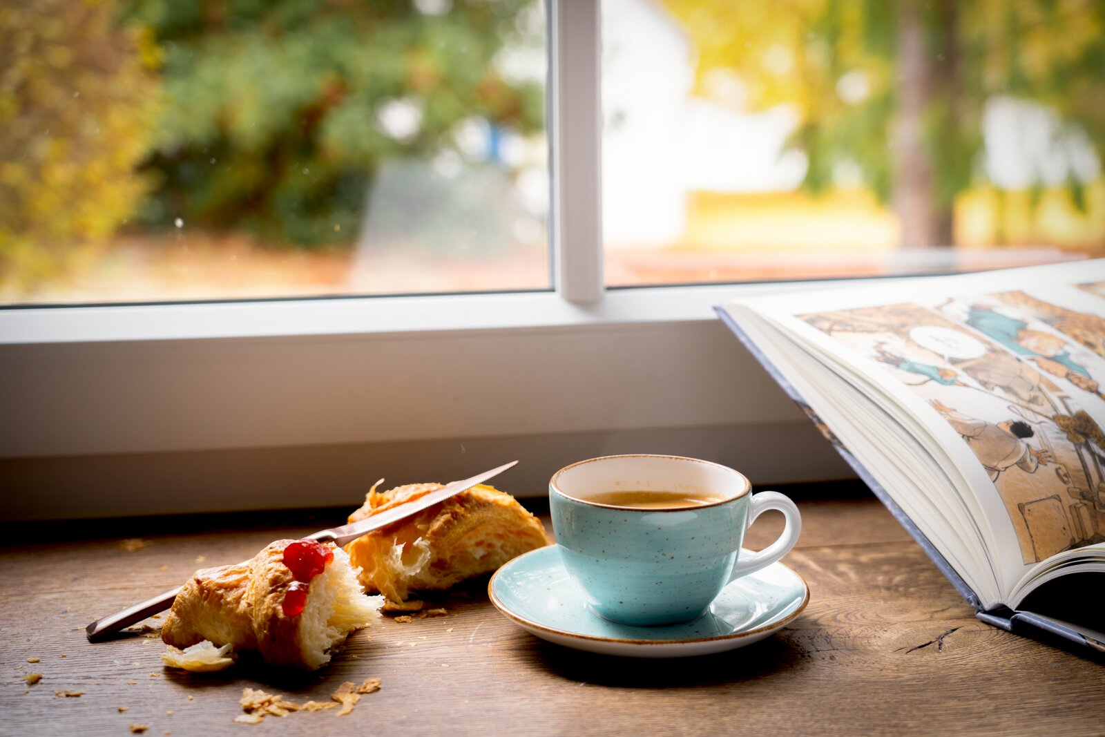 Hellblaue Kaffeetasse mit Untertasse auf Holztisch am Fenster, daneben Croissant mit Marmelade und aufgeschlagenes Buch; Perspektive: leicht von vorne auf Tischhöhe