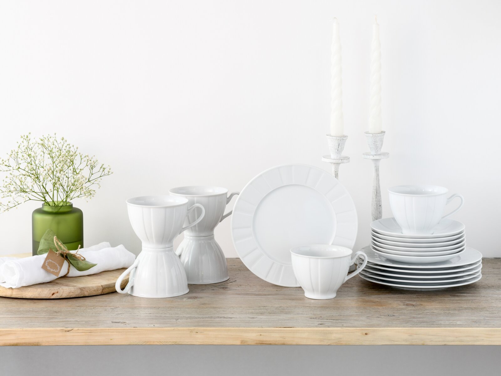 Weißes Porzellan-Geschirrset mit Tassen, Tellern und Untertassen auf Holztisch dekoriert mit Kerzenständern und Vase, frontal in leichter Schrägansicht