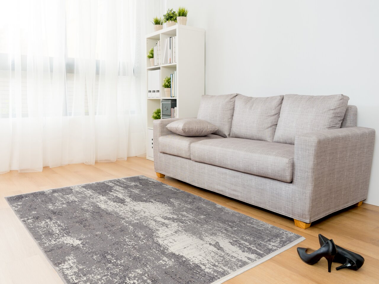 Moderner Designer Teppich in Grau- und Weißtönen auf hellem Holzboden, vor einem grauen Sofa in einem hellen Wohnzimmer mit großen Fenstern, seitliche Perspektive.