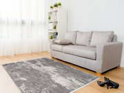 Moderner Designer Teppich in Grau- und Weißtönen auf hellem Holzboden, vor einem grauen Sofa in einem hellen Wohnzimmer mit großen Fenstern, seitliche Perspektive.