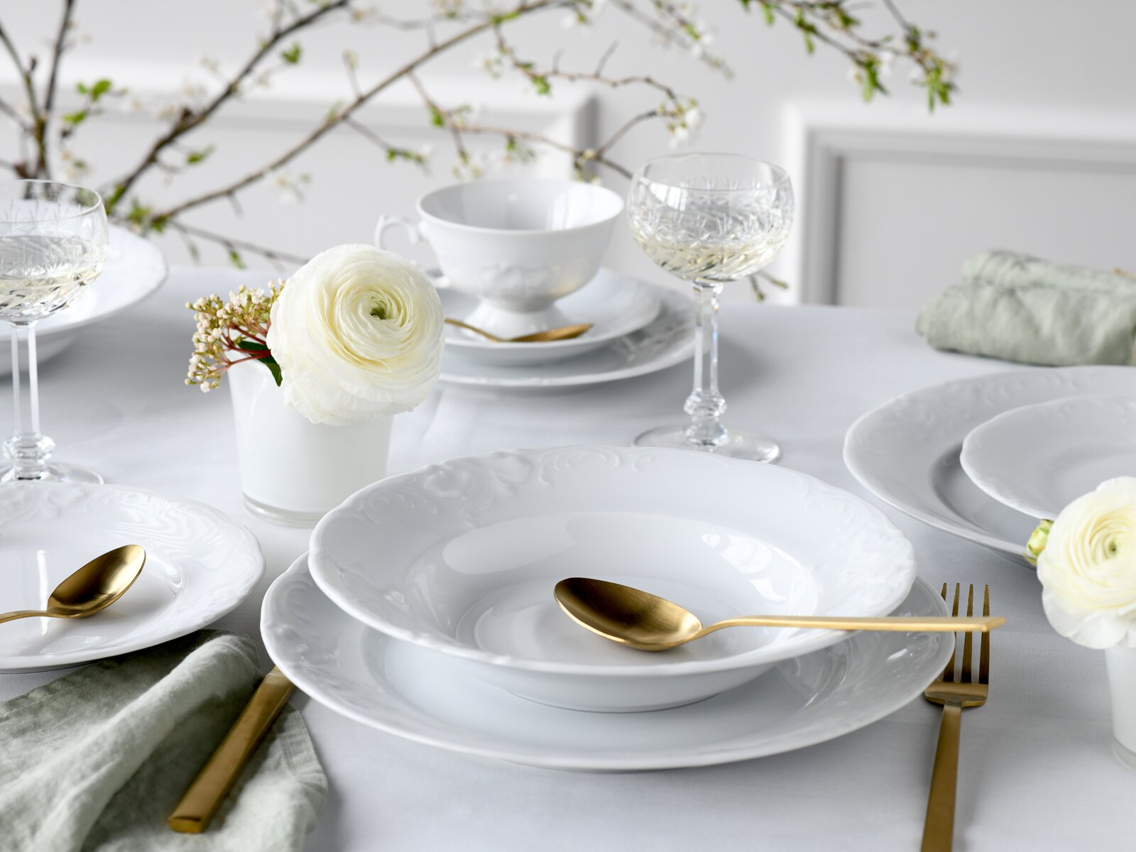 Elegantes weißes Porzellan-Geschirrset mit geprägtem Rand, goldfarbenem Besteck und Kristall-Weingläsern auf gedecktem Tisch mit weißen Blumen, aufgenommen in leicht erhöhter schräger Perspektive von vorn.