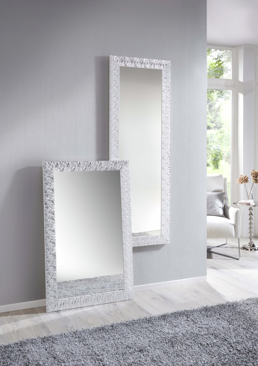 Zwei rechteckige Spiegel mit dekorativem Rahmen, einer stehend an der Wand und der andere an die Wand gelehnt, in einem modernen, hellen Raum mit Teppich und Fenster im Hintergrund.