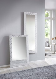 Zwei rechteckige Spiegel mit dekorativem Rahmen, einer stehend an der Wand und der andere an die Wand gelehnt, in einem modernen, hellen Raum mit Teppich und Fenster im Hintergrund.