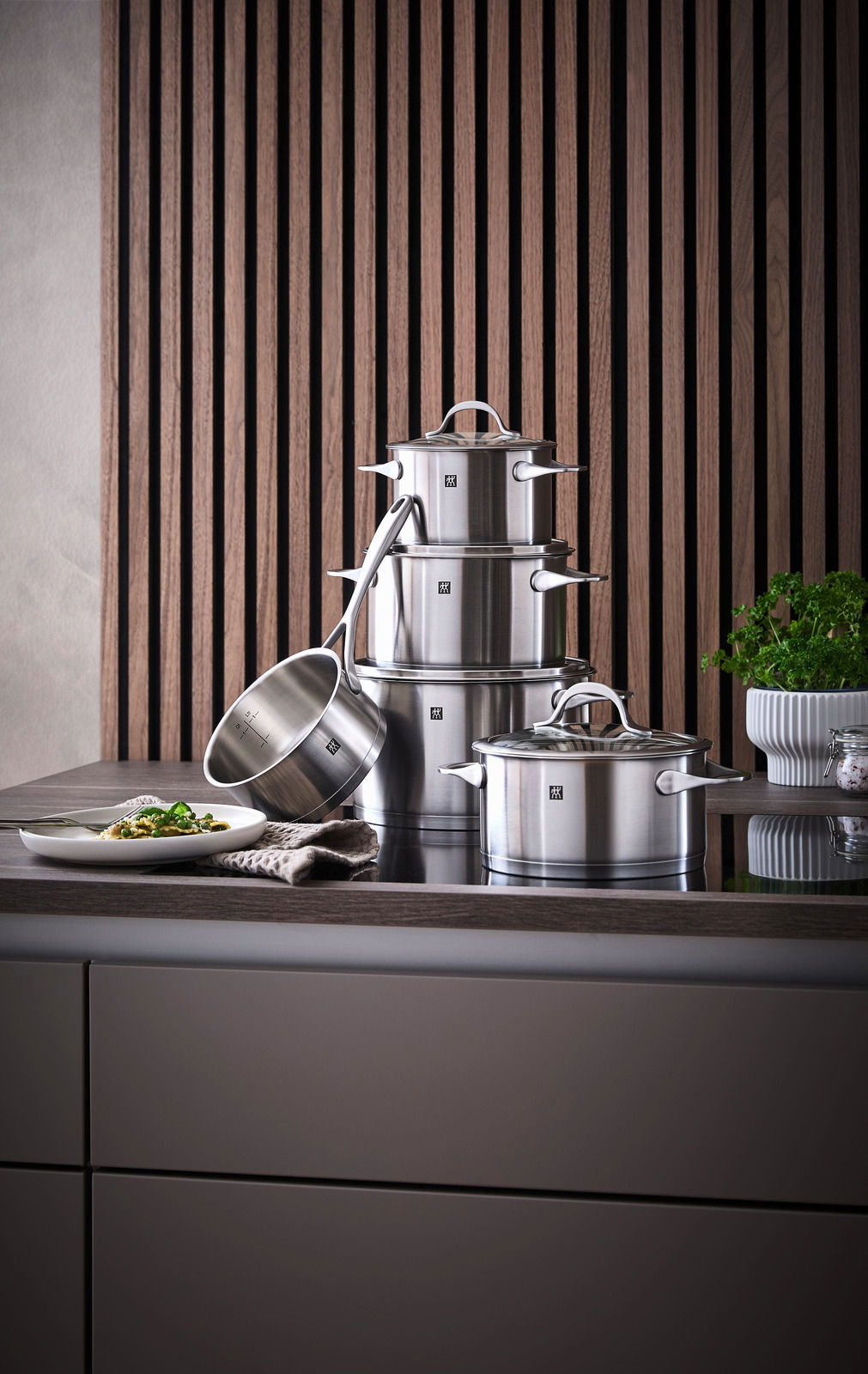 ZWILLING Topfset 5-tlg. ESSENCE Topfset Essence 5-tlg. aus Edelstahl, bestehend aus mehreren Töpfen und einer Stielkasserolle, auf einer Küchenarbeitsplatte vor einer Holzvertäfelung präsentiert. Perspektive von vorne.