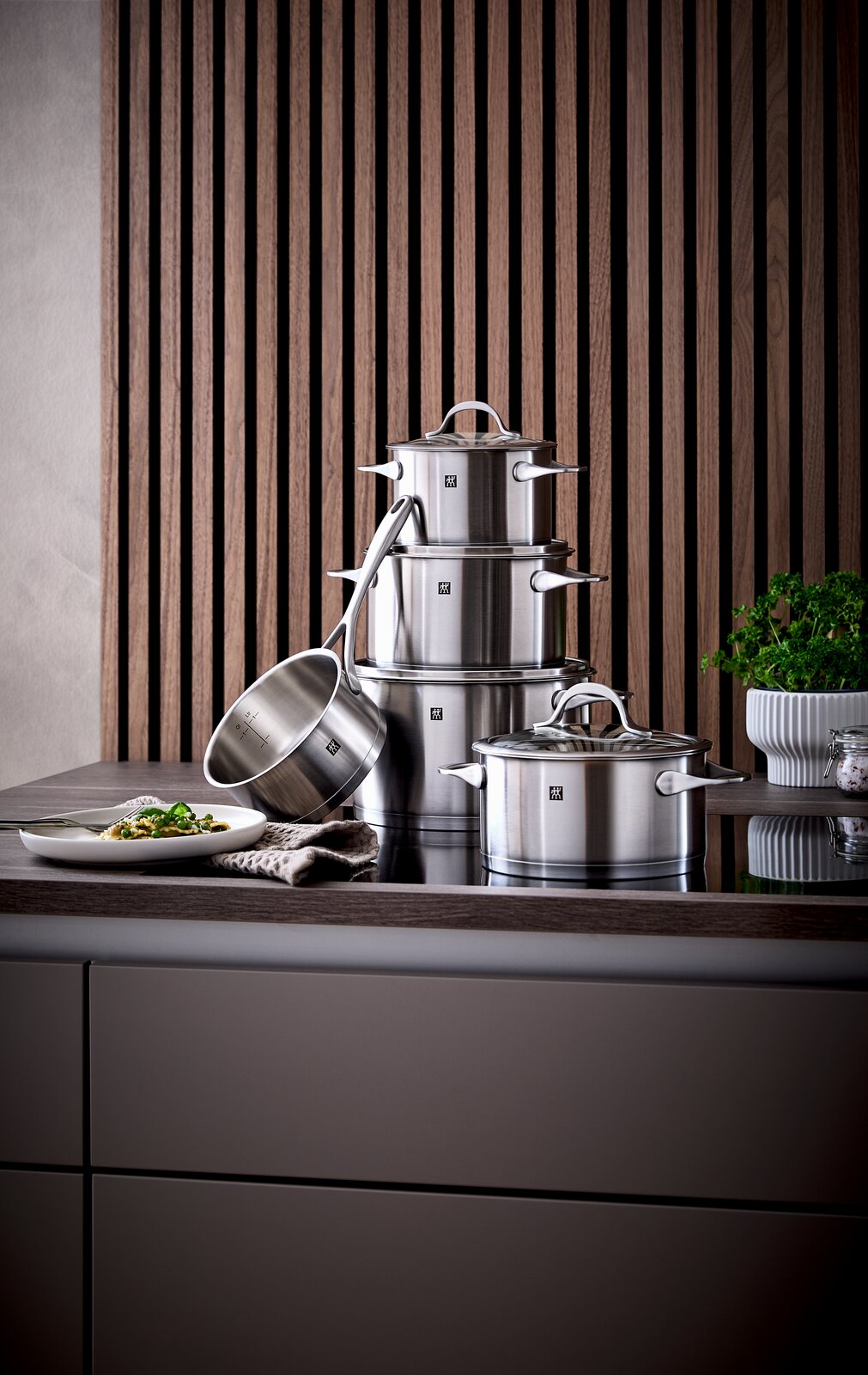 ZWILLING Topfset 5-tlg. ESSENCE Topfset Essence 5-tlg. aus Edelstahl, bestehend aus mehreren Töpfen und einer Stielkasserolle, auf einer Küchenarbeitsplatte vor einer Holzvertäfelung präsentiert. Perspektive von vorne.