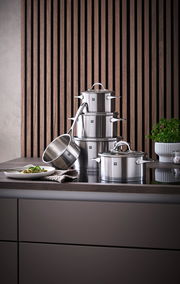 Topfset Essence 5-tlg. aus Edelstahl, bestehend aus mehreren Töpfen und einer Stielkasserolle, auf einer Küchenarbeitsplatte vor einer Holzvertäfelung präsentiert. Perspektive von vorne.